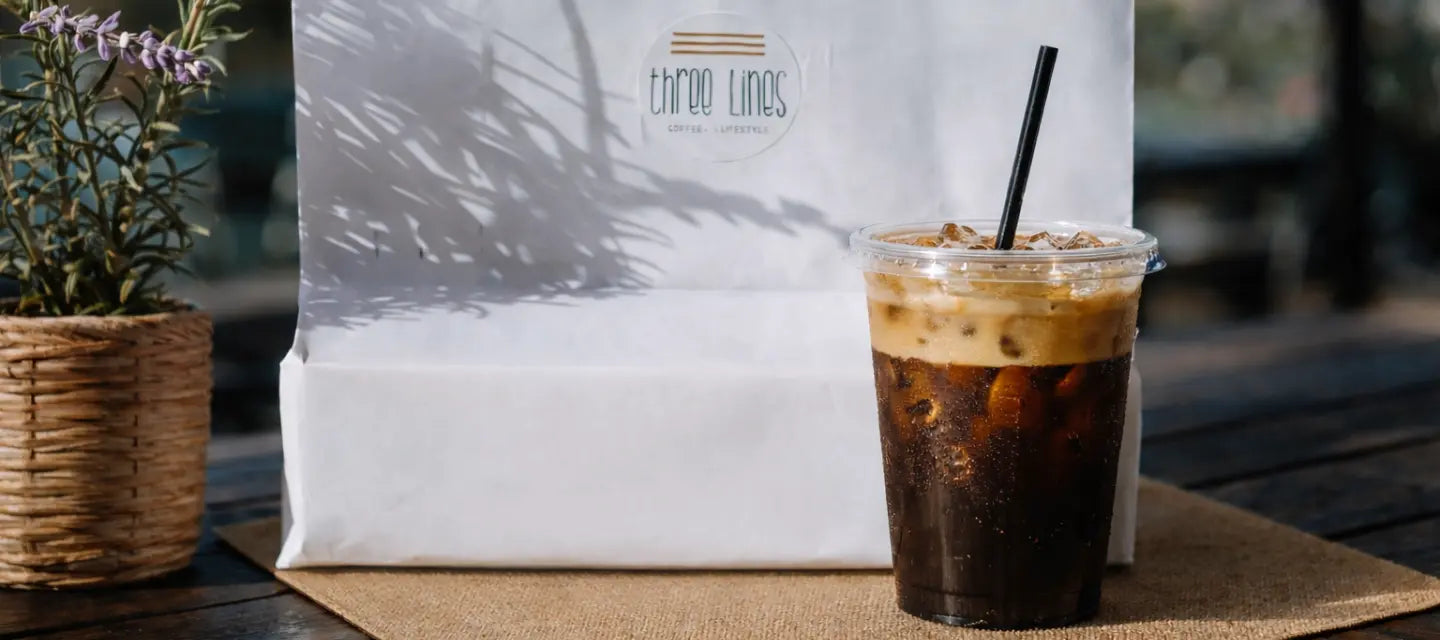 Cold brew coffee in a clear plastic cup with a black straw on a wooden surface, with a white bag labeled 'ThreeLinesCoffee' in the background.